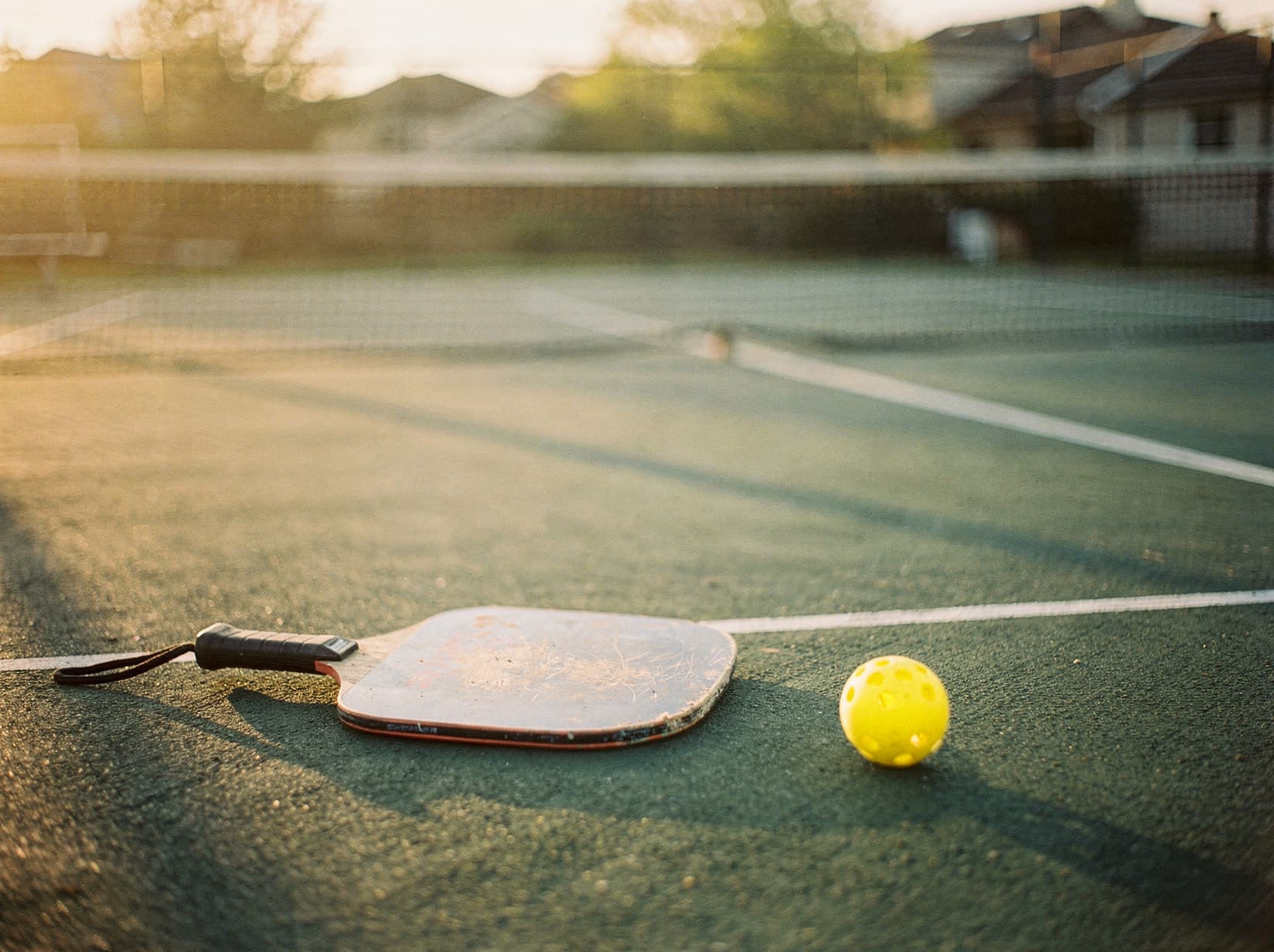 A pickleball paddle and ball resting on a court at golden hour